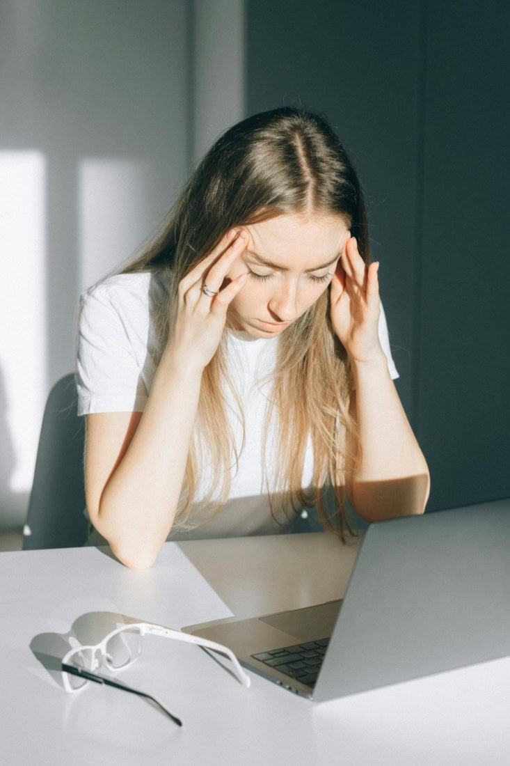Huilende vrouw met hoofdpijn, werkstress en vermoeidheid, laptop en glas op tafel.
