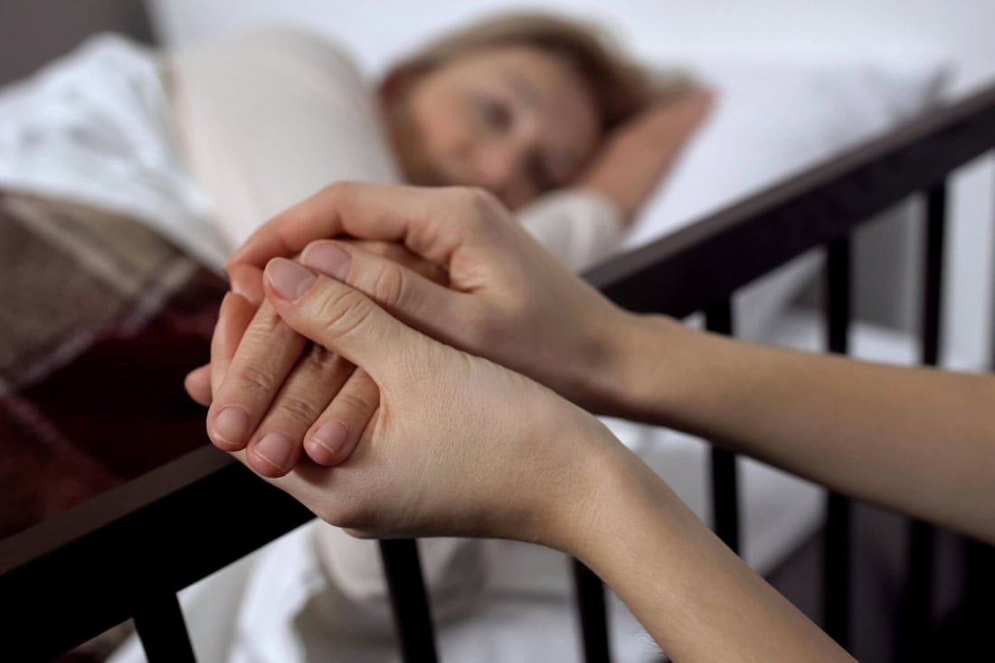 Zorgzame hand vasthoudt hand van zieke persoon in bed, symboliseert steun en compassie.